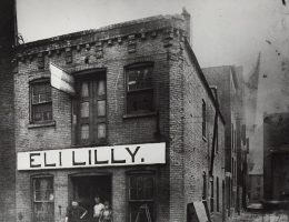 Zdjęcie pierwszego budynku przedsiębiorstwa. Eli Lilly z synem Josiahem stoją po lewej stronie drzwi
